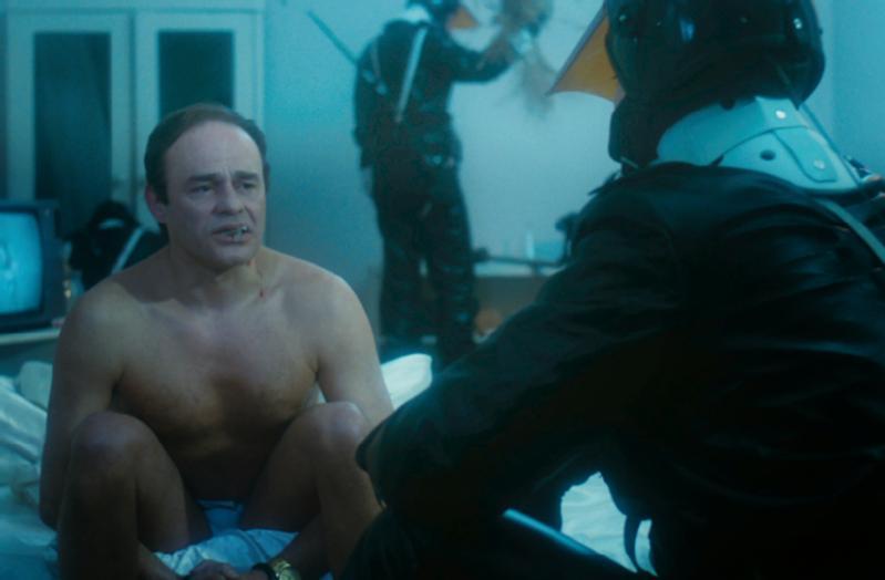 Mężczyzna z gołą klatką piersiową siedzi na łóżku, wygląda na zaniepokojonego, naprzeciwko osoby w czarnym taktycznym rynsztunku i hełmie. W tle dwie inne postacie w podobnym rynsztunku powstrzymują kogoś. Scena jest oświetlona niebieskawym odcieniem.