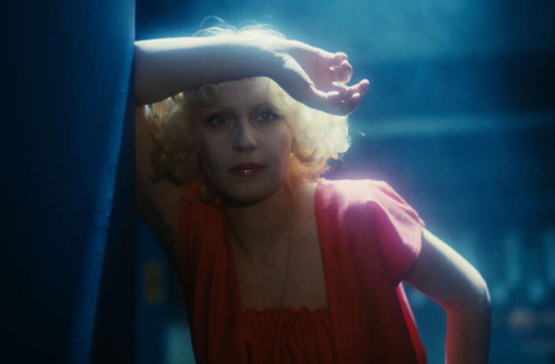 Kobieta z krótkimi, kręconymi blond włosami i czerwoną szminką opiera się o powierzchnię, jej ramię spoczywa na czole. Ma na sobie czerwony top i jest oświetlona miękkim, niebieskim światłem.