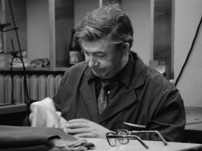 Mężczyzna w roboczym płaszczu siedzi przy stole, czyszcząc lub sprawdzając przedmiot szmatką. Okulary leżą na stole przed nim. W tle widać półki i różne przedmioty.