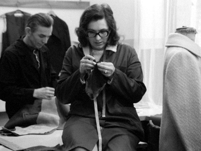 Kobieta o ciemnych włosach i okularach szyje tkaninę ręcznie, siedząc przy stole warsztatowym; mężczyzna pracuje w tle. Oboje wydają się skupieni, a otoczenie przypomina warsztat krawiecki.