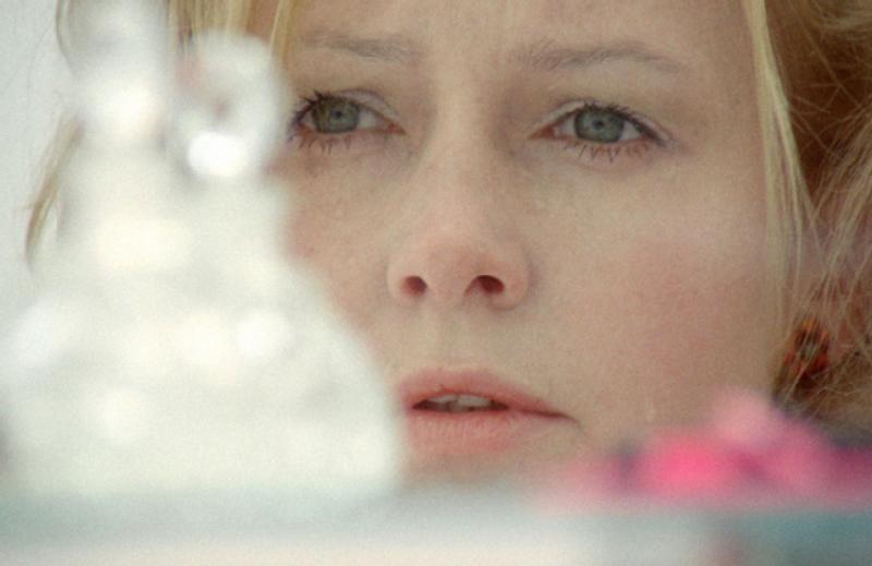 Zbliżenie kobiety o blond włosach i jasnych oczach, zamyślonej i patrzącej przed siebie. Na pierwszym planie widać rozmazane kształty i obiekty nieostre.