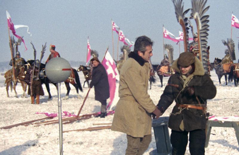 Dwóch mężczyzn stoi i ściska sobie dłonie na zaśnieżonym polu, jeden z nich ubrany jest w historyczną zbroję i skrzydła. Za nimi są ludzie na koniach i wiele różowo-białych flag. Scena wydaje się być częścią planu filmowego lub rekonstrukcji.