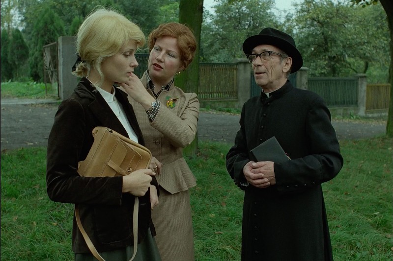 Młoda kobieta z brązową torbą stoi w trawiastym parku, podczas gdy starsza kobieta poprawia jej kołnierzyk. Mężczyzna ubrany w czarny strój duchowny i kapelusz stoi w pobliżu, trzymając książkę. W tle widać drzewa i drewniany płot.