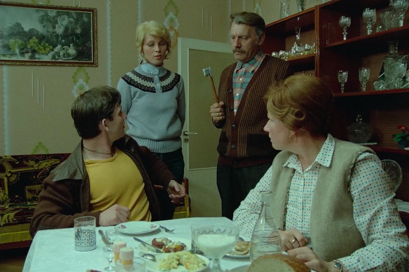 Cztery osoby siedzą przy stole jadalnym zastawionym jedzeniem i napojami. Starszy mężczyzna stoi trzymając małą siekierę, podczas gdy pozostali trzej, dwie kobiety i mężczyzna, patrzą na niego z niepokojem lub zdziwieniem. Pokój ma wystrój w stylu vintage.