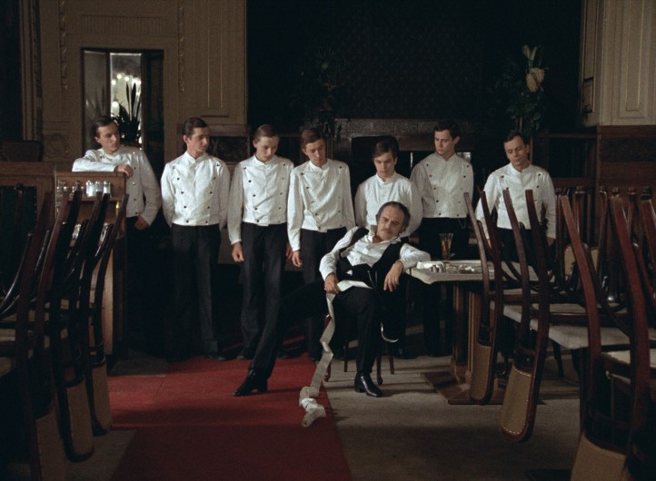 Siedmiu kelnerów w białych uniformach stoi w szeregu i patrzy na siedzącego mężczyznę w czarno-białym stroju formalnym, który wygląda na zmęczonego i zaniedbanego, garbiącego się przy stoliku w niemal pustej, eleganckiej restauracji.