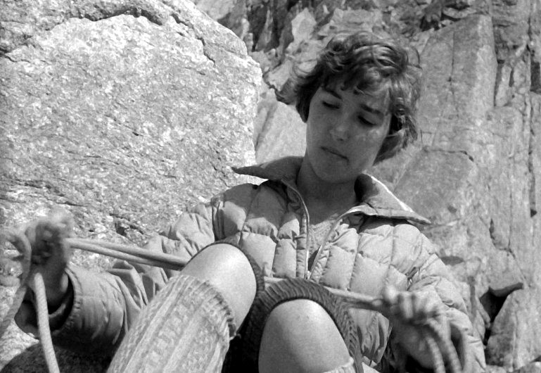 Młoda kobieta ubrana w pikowaną kurtkę i sięgające do kolan skarpetki obsługuje linę wspinaczkową, siedząc blisko skalistej powierzchni i patrząc w dół ze skupionym wyrazem twarzy.