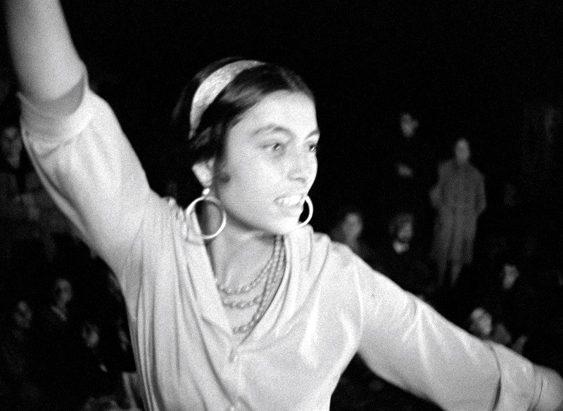 Młoda kobieta z kolczykami w kształcie kółek i opaską na głowę podnosi rękę, lekko się uśmiechając, a na tle słabo oświetlonych osób widać rozmazane postacie. Obraz jest czarno-biały.
