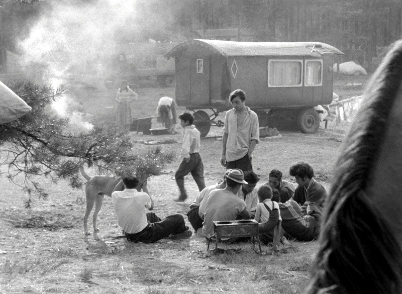 Grupa ludzi siedzi w kręgu na ziemi na zewnątrz, w pobliżu unosi się dym. Obok nich stoi pies. W tle widać wóz i kilka osób wśród drzew w wiejskim otoczeniu. Obraz jest czarno-biały.