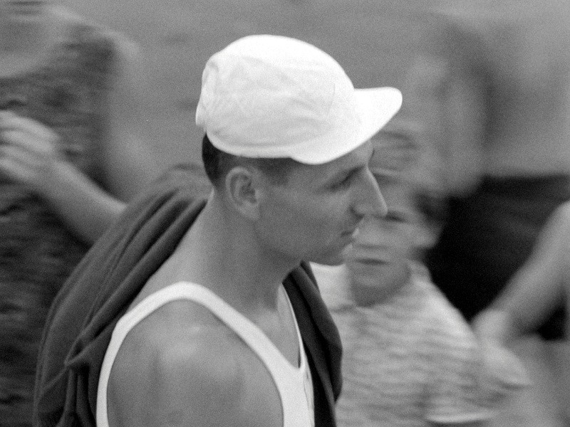 Mężczyzna w białej czapce i podkoszulku widoczny jest z profilu, niosąc ciemną kurtkę na ramieniu. W tle widać niewyraźne postacie, co sugeruje swobodne otoczenie na zewnątrz.