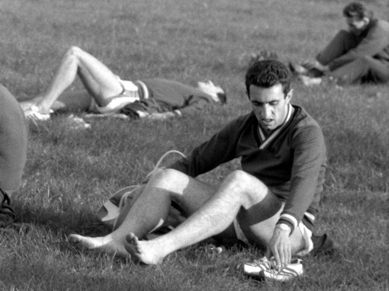 Mężczyzna siedzi na trawie, patrzy w dół, wiążąc but, ubrany w strój sportowy. W tle dwie inne osoby w podobnych ubraniach leżą na trawie, prawdopodobnie odpoczywając lub się rozciągając.