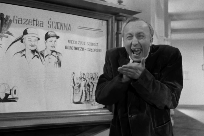 Mężczyzna w garniturze śmieje się radośnie, trzymając dłonie w dłoniach, stojąc obok zawieszonej na ścianie tablicy z ilustracjami i tekstem w języku polskim w czarno-białym otoczeniu.