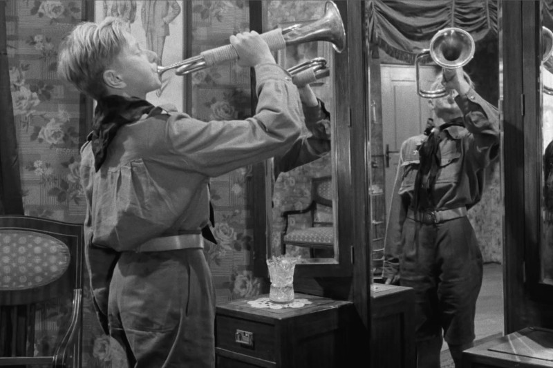 Młody chłopiec w mundurze dmucha w trąbkę przed lustrem, odbijając swój obraz. W pokoju jest wzorzysta tapeta, krzesło i toaletka ze szklanym pojemnikiem.