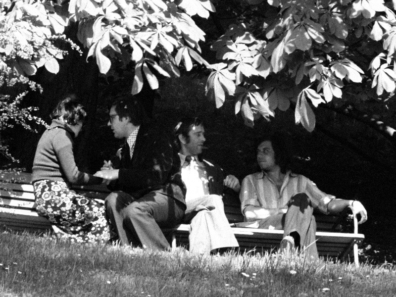 Cztery osoby siedzą i rozmawiają na ławce pod drzewem, a światło słoneczne prześwituje przez liście. Dwie osoby stoją naprzeciwko siebie, podczas gdy pozostali wyglądają na zrelaksowanych, co sugeruje swobodną rozmowę. Scena wydaje się pochodzić z lat 70.