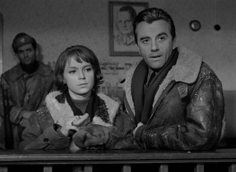 Młoda kobieta i mężczyzna w futrzanych płaszczach stoją przy ladzie, poważnie patrząc przed siebie. Inny mężczyzna w mundurze stoi w tle, a portret wisi na ścianie za nimi w czarno-białej scenie.