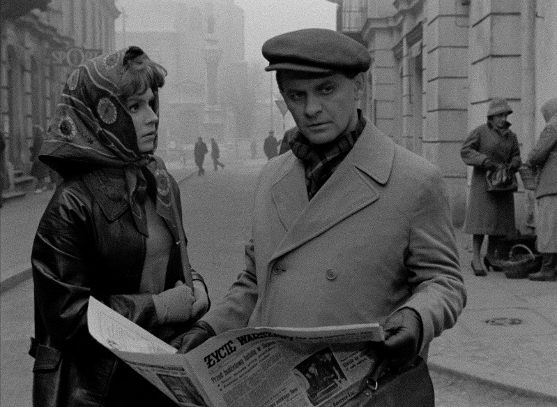 Kobieta w chuście na głowie i mężczyzna w płaszczu i czapce stoją na miejskiej ulicy; mężczyzna trzyma gazetę. Scena jest czarno-biała, z rozmazanymi pieszymi i budynkami w tle.