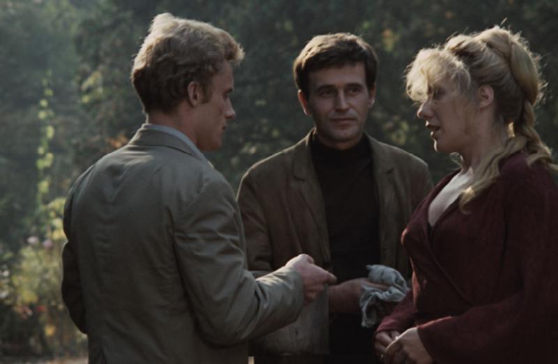 Trzy osoby stoją na zewnątrz i rozmawiają. Jeden mężczyzna stoi naprzeciwko kobiety, podając jej coś, podczas gdy inny mężczyzna stoi między nimi i patrzy. W tle widać drzewa i łagodne światło słoneczne.