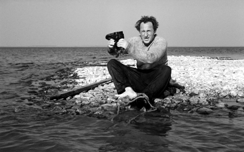 Mężczyzna siedzi boso na skalistym brzegu, trzymając parę butów, a woda chlapie wokół jego stóp. Morze i horyzont są widoczne w tle pod czystym niebem.