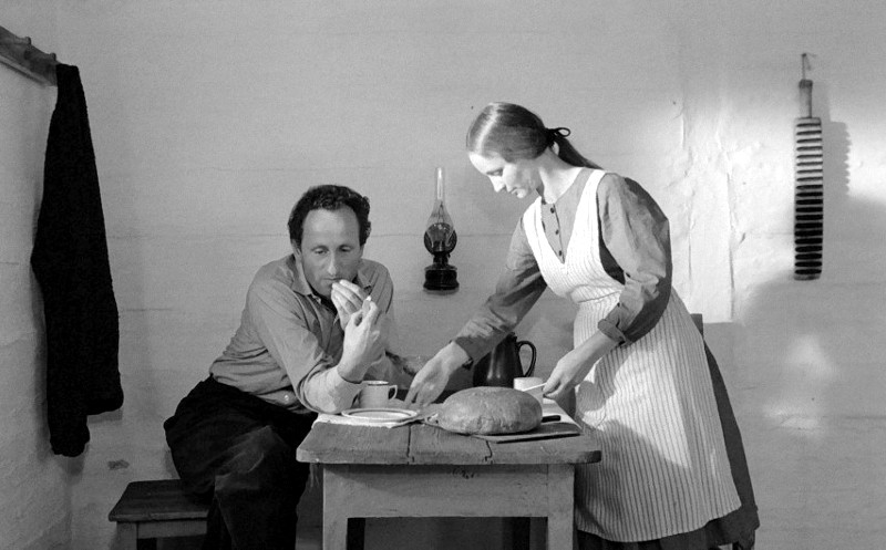 Mężczyzna siedzi przy małym drewnianym stole, jedząc chleb, podczas gdy kobieta w fartuchu stoi obok niego, uśmiecha się i serwuje jedzenie. Scena jest prosta, z bochenkiem chleba, kubkami i lampą na stole, na tle prostej białej ściany.
