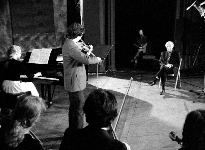 Czarno-białe zdjęcie przedstawiające skrzypka grającego na scenie z pianistą i innymi muzykami, podczas gdy starszy mężczyzna siedzi na krześle naprzeciwko nich; w tle siedzi jeszcze jedna osoba.