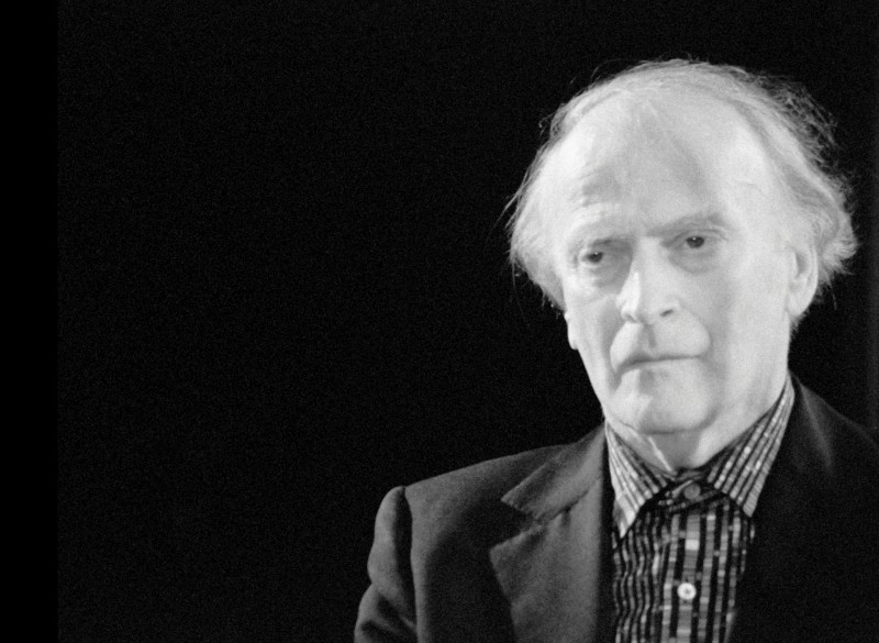 Czarno-białe zdjęcie starszego mężczyzny o jasnych włosach, ubranego w ciemną kurtkę i kraciastą koszulę, patrzącego lekko w bok na jednolitym ciemnym tle.