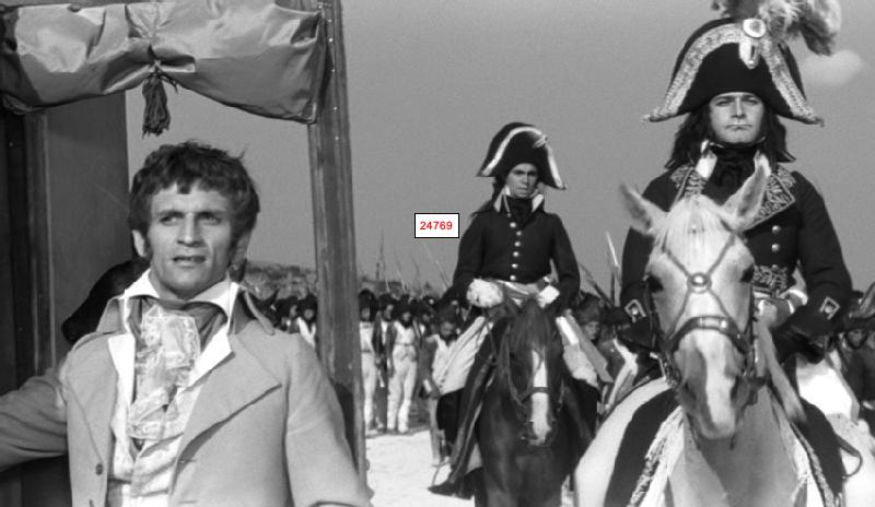 Mężczyzna w stroju z epoki stoi na pierwszym planie, a za nim dwóch mężczyzn w napoleońskich mundurach wojskowych jedzie konno, prowadząc formację żołnierzy niosących karabiny. Scena jest czarno-biała.