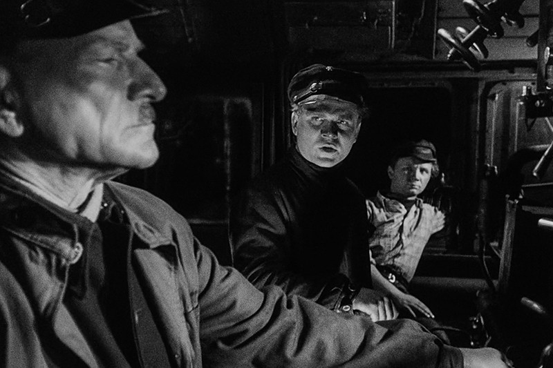 Trzech mężczyzn w ciemnych mundurach roboczych i czapkach siedzi w słabo oświetlonej kabinie pociągu, jeden mężczyzna na pierwszym planie skupia się na sterowaniu, a dwóch innych uważnie na niego patrzy. Scena jest czarno-biała.