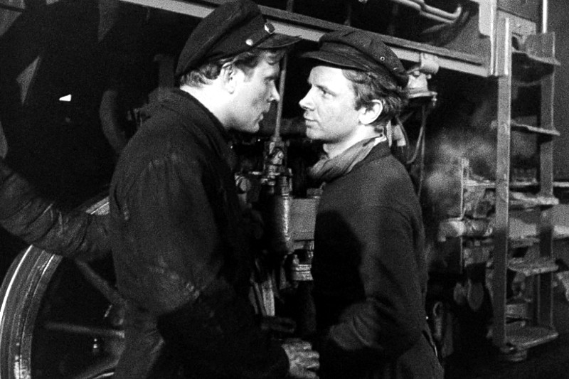 Dwóch mężczyzn w mundurach roboczych i czapkach stoi twarzą w twarz przed dużą lokomotywą, wyglądając poważnie i pogrążonych w napiętej rozmowie. Scena jest czarno-biała.