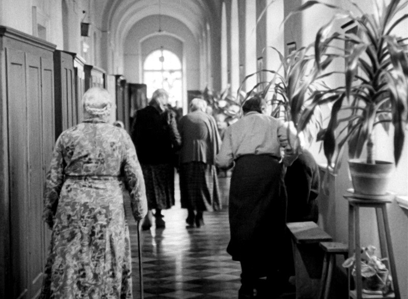 Starsi ludzie idą długim, łukowatym korytarzem z kafelkową podłogą i doniczkowymi roślinami wzdłuż ścian, niektórzy używają lasek jako podpórki. Scena ma czarno-biały, zabytkowy wygląd.
