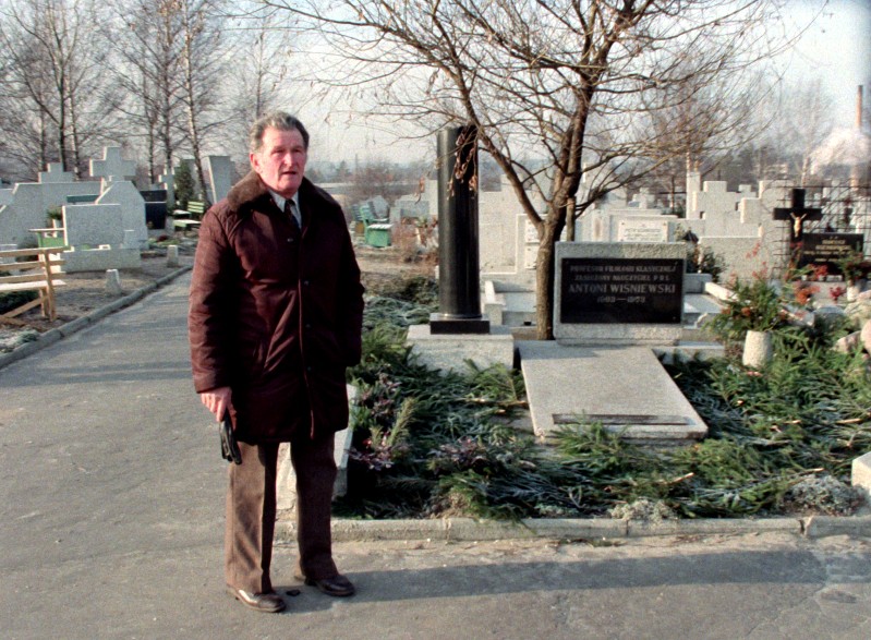 Starszy mężczyzna w brązowym płaszczu stoi, trzymając kapelusz, przy grobie na cmentarzu, otoczony nagrobkami i bezlistnymi drzewami, w zimny, pochmurny dzień.