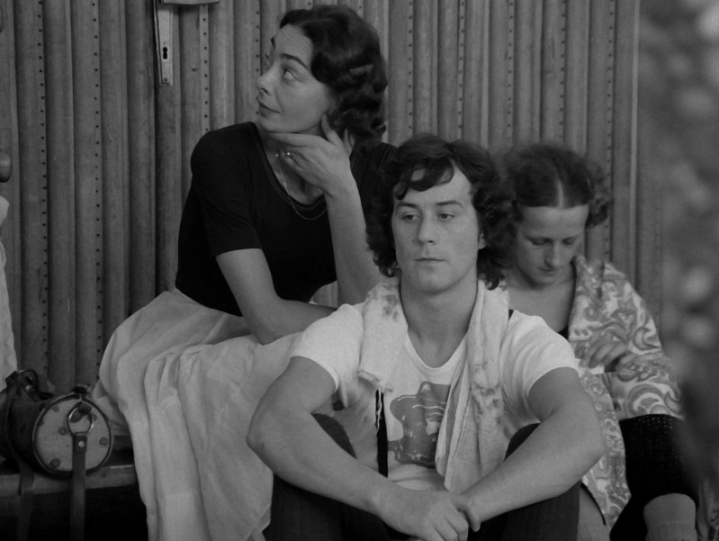 Trzy osoby siedzą blisko siebie w pomieszczeniu. Kobieta w ciemnym topie i jasnej spódnicy patrzy w bok, mężczyzna z kręconymi włosami siedzi z przodu, a inna kobieta patrzy w dół, częściowo przykryta kocem. Nastrój wydaje się kontemplacyjny.