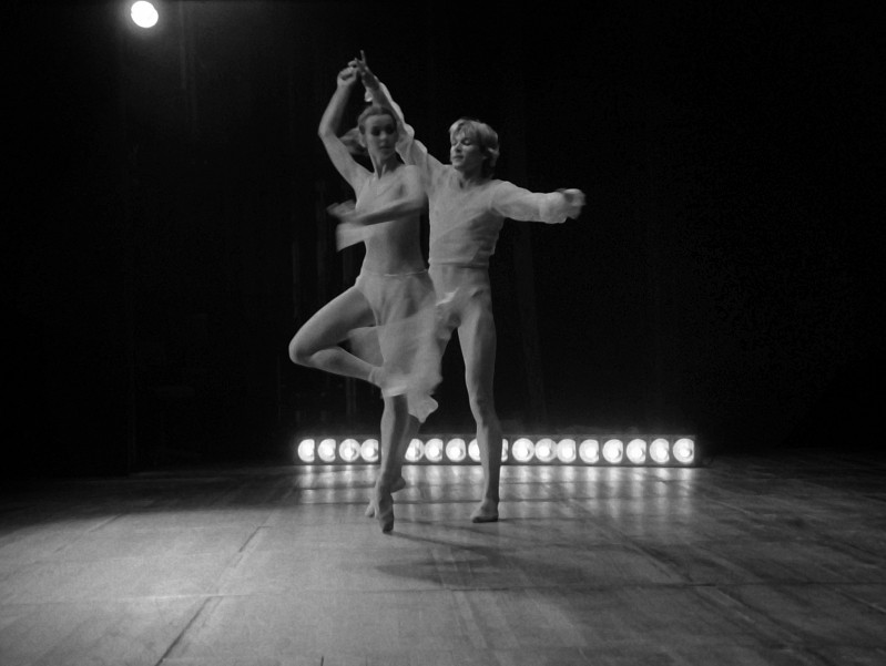 Dwie tancerki w prześwitujących, zwiewnych strojach występują na słabo oświetlonej scenie z rzędem jasnych świateł za nimi. Jedna tancerka jest w połowie obrotu z uniesioną nogą, podczas gdy druga porusza się z wyciągniętymi ramionami.