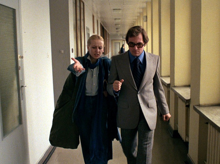 Kobieta w niebieskim stroju gestykuluje, idąc obok mężczyzny w szarym garniturze i ciemnych okularach przeciwsłonecznych wzdłuż jasno oświetlonego korytarza z dużymi oknami.