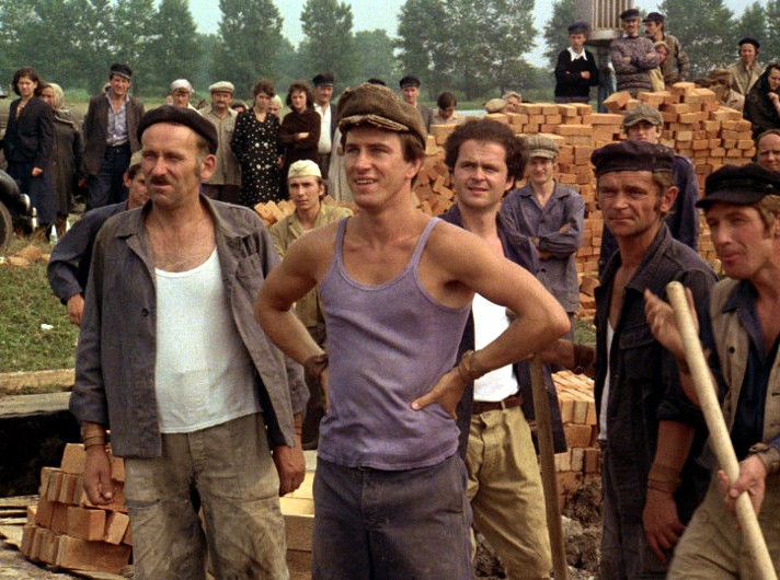 Grupa mężczyzn w ubraniach roboczych i czapkach stoi obok ułożonych w stosy cegieł na placu budowy. W tle widać ludzi, a w oddali widać drzewa.