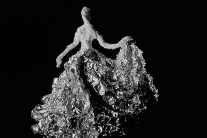 Postać w dramatycznej, odblaskowej sukni z pogniecionego, metalicznego materiału stoi na czarnym tle, trzymając część sukni na bok. Górna część ciała i głowa są gładkie i białe.