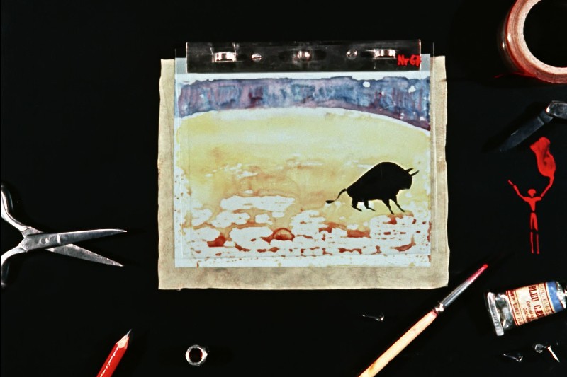 Rysunek byka na pergaminie umieszczony jest na czarnej powierzchni. Wokół niego znajdują się przybory artystyczne, m.in. nożyczki, ołówek, pędzel, rolka taśmy, tubka czerwonej farby i mała butelka.