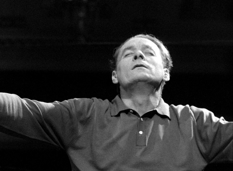 Mężczyzna w koszuli z kołnierzykiem stoi z wyciągniętymi przed siebie ramionami i zamkniętymi oczami, sprawia wrażenie, jakby był głęboko pogrążony w myślach lub emocjach, na ciemnym tle.