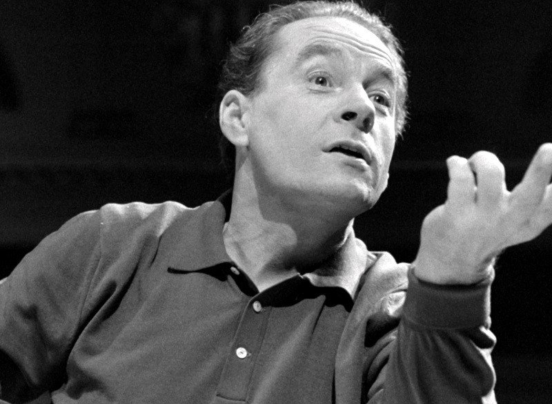 Mężczyzna z zaczesanymi do tyłu włosami, ubrany w koszulę z kołnierzykiem, gestykuluje ekspresyjnie ręką, patrząc lekko w górę z wyrazem zaangażowania. Zdjęcie jest czarno-białe.