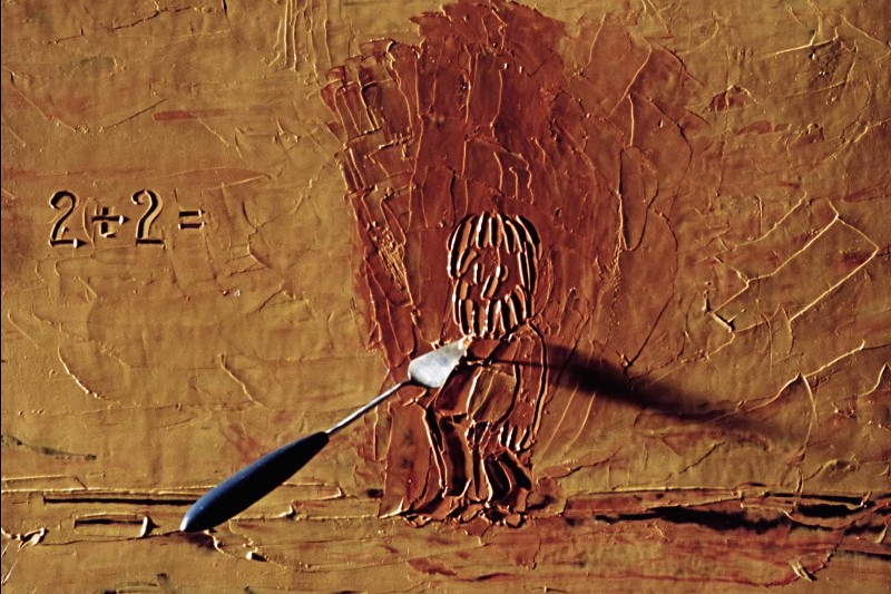 Widelec spoczywa na teksturowanej brązowej powierzchni z wyrzeźbioną postacią osoby i wytrawionym obok niej równaniem 2+2=. Cień widelca pada na postać i tło.