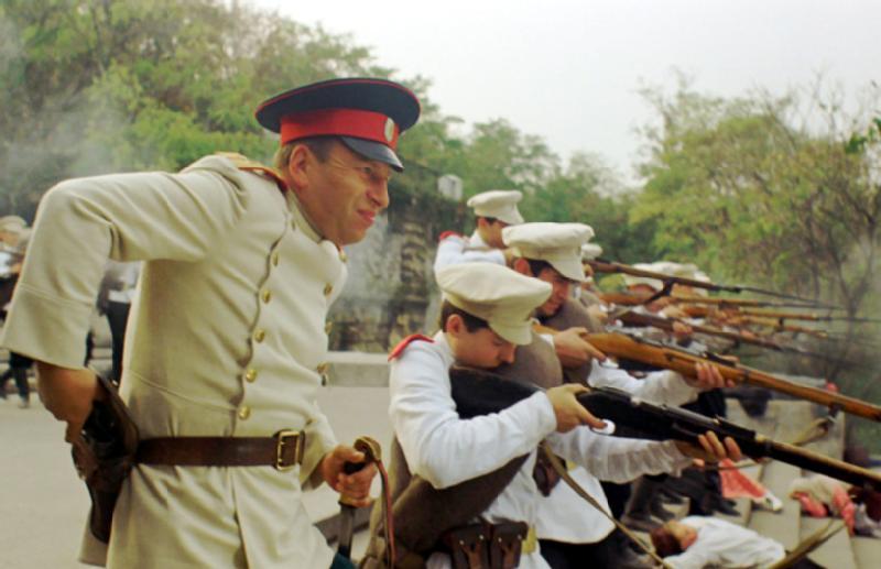 Grupa mężczyzn ubranych w mundury wojskowe z początku XX wieku celuje z karabinów w szyku na zewnątrz, z jednym oficerem w białym płaszczu i czerwonej czapce stojącym na czele. W tle widać drzewa i dym.