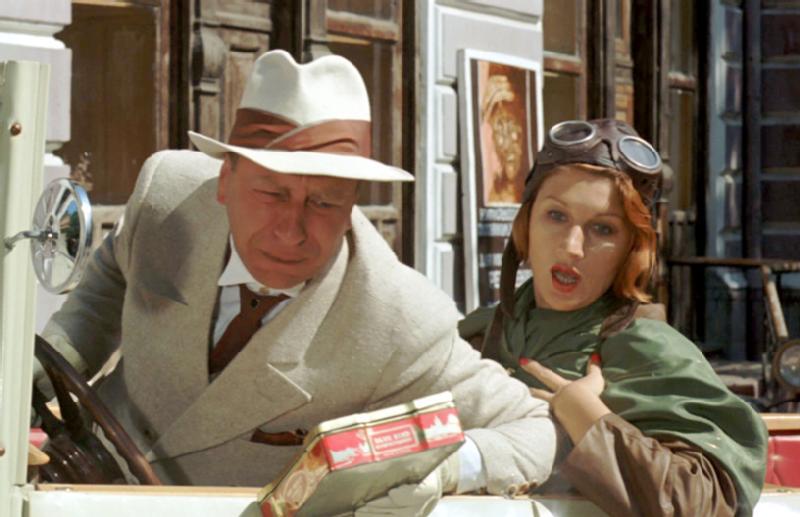 Mężczyzna w jasnym garniturze i kapeluszu siedzi w zabytkowym samochodzie, pochylony do przodu, a obok niego siedzi zaskoczona kobieta w zielonym płaszczu i goglach lotniczych, trzymająca paczkę herbaty, a w tle widać budynki.