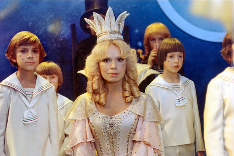 Kobieta w błyszczącej koronie i ozdobnej sukni stoi przed grupą dzieci ubranych w białe marynarskie stroje, na niebieskim tle z dużym kołem.