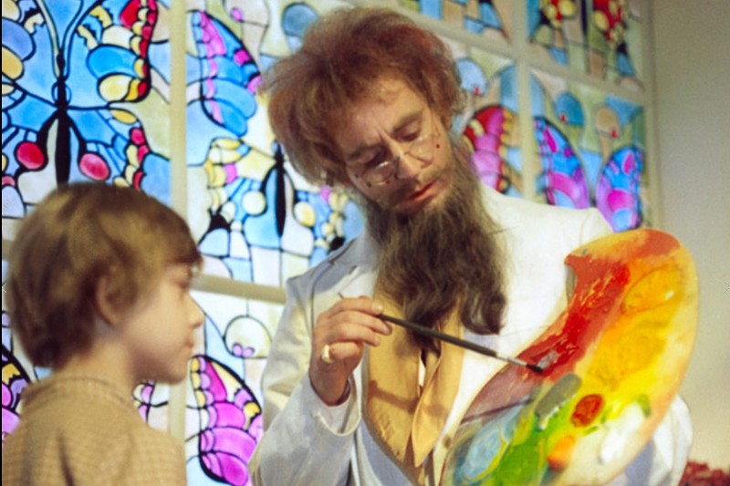 Mężczyzna z długą brodą i okularami, ubrany na biało, trzyma kolorową paletę malarską i pędzel, podczas gdy małe dziecko stoi w pobliżu. Za nimi znajdują się witraże z motywami motyli.