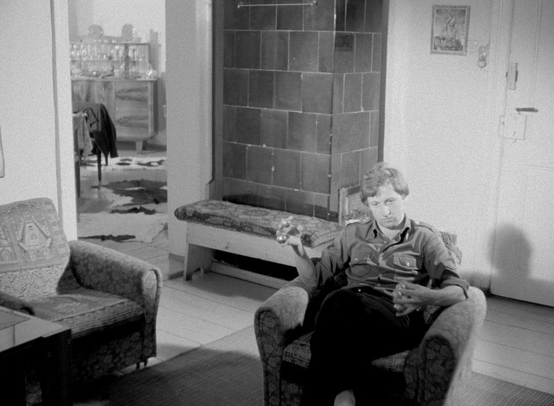 Osoba siedzi sama w fotelu w skromnie umeblowanym pokoju, trzymając drinka. W pokoju znajduje się kafelkowy kominek, wzorzyste krzesła i otwarte drzwi odsłaniające inny pokój w tle. Scena jest czarno-biała.