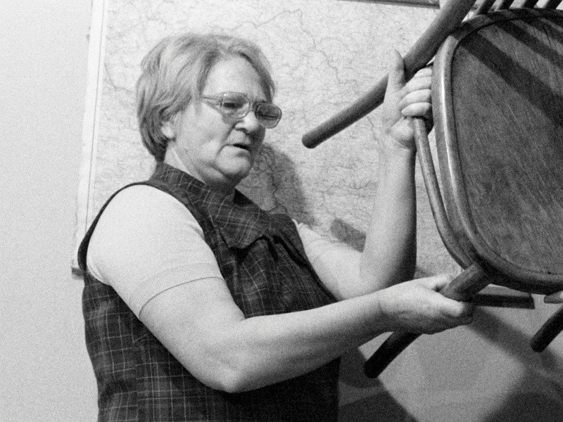 Starsza kobieta w okularach podnosi drewniane krzesło przed wiszącą na ścianie mapą. Obraz jest czarno-biały.