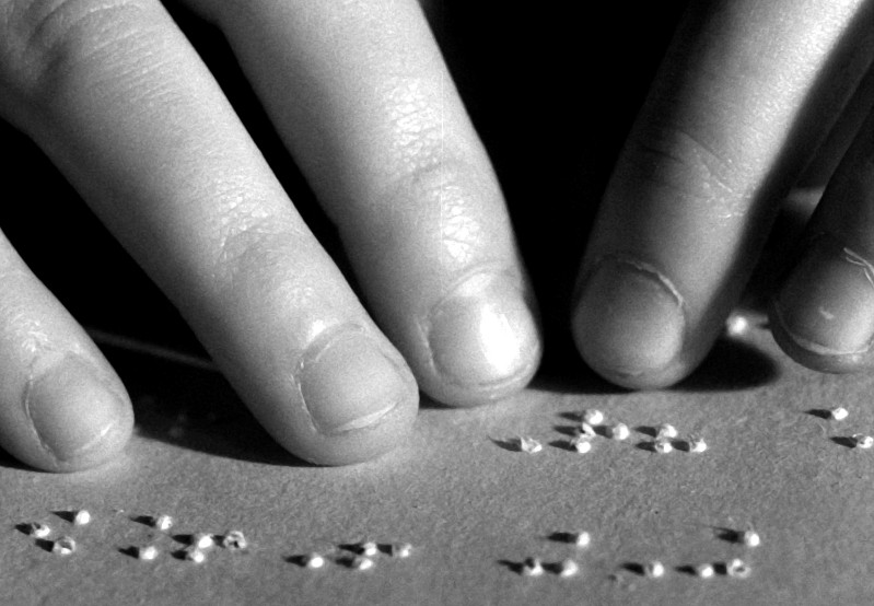 Zbliżenie dwóch dłoni, których palce delikatnie się dotykają i czytają wypukłe kropki Braille’a na kartce papieru.