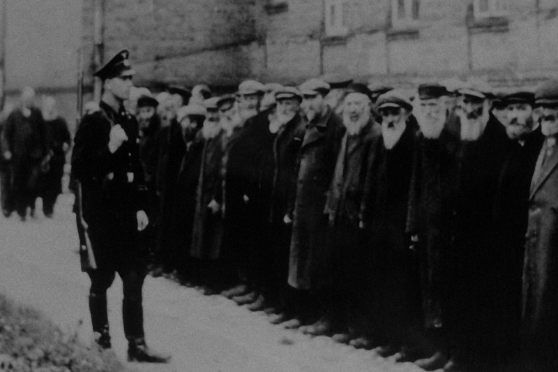 Czarno-białe historyczne zdjęcie przedstawia umundurowanego oficera stojącego przed szeregiem ponurych mężczyzn ubranych w płaszcze i czapki, prawdopodobnie z początku lub połowy XX wieku, na tle ceglanego budynku.