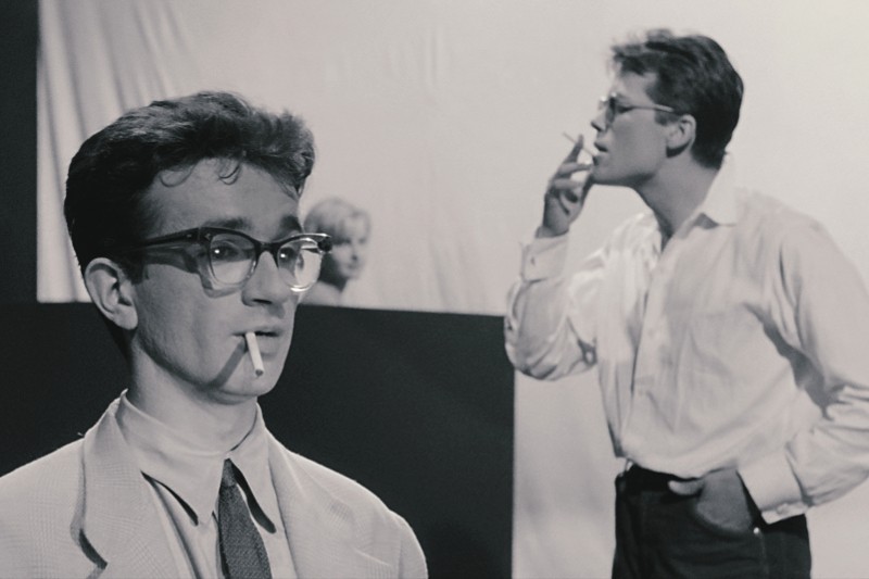Dwóch młodych mężczyzn w okularach i koszulach stoi w pomieszczeniu, każdy z papierosem w ustach. Jeden jest na pierwszym planie, patrzy przed siebie, a drugi jest w tle, wydychając dym. Obraz jest czarno-biały.