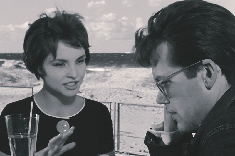 Kobieta z krótkimi włosami ożywiająco rozmawia z mężczyzną w okularach, który wydaje się pogrążony w myślach. Siedzą przy stole ze szklanką wody, a w tle widać plażę z płotem.