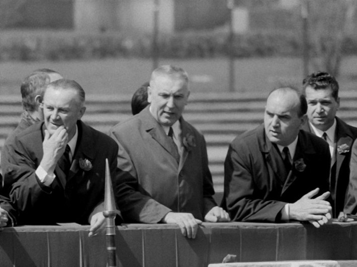 Grupa czterech mężczyzn w garniturach stoi na zewnątrz za barierką, rozmawiając. Obraz jest czarno-biały, a scena wydaje się być formalna lub oficjalna.