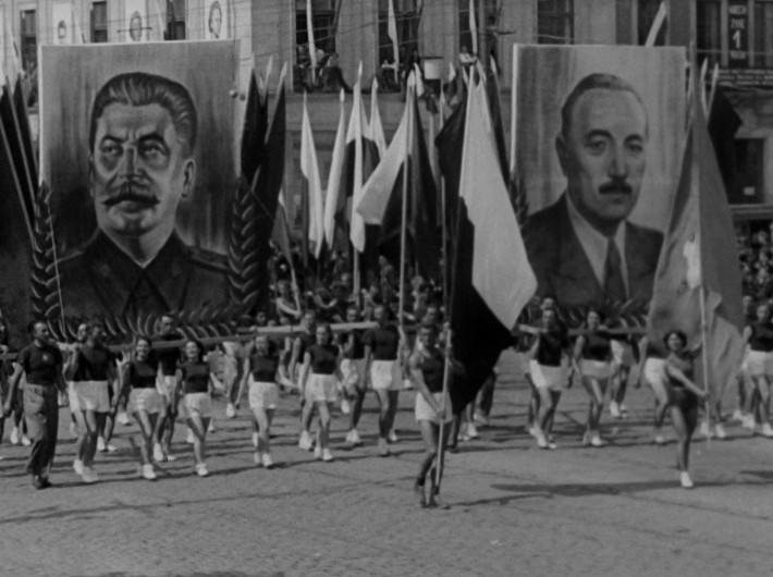 Parada ludzi ubranych w krótkie spodenki i ciemne koszule, niosących duże flagi i dwa ogromne portrety mężczyzn z wąsami, eksponowane w widocznym miejscu na miejskim placu.